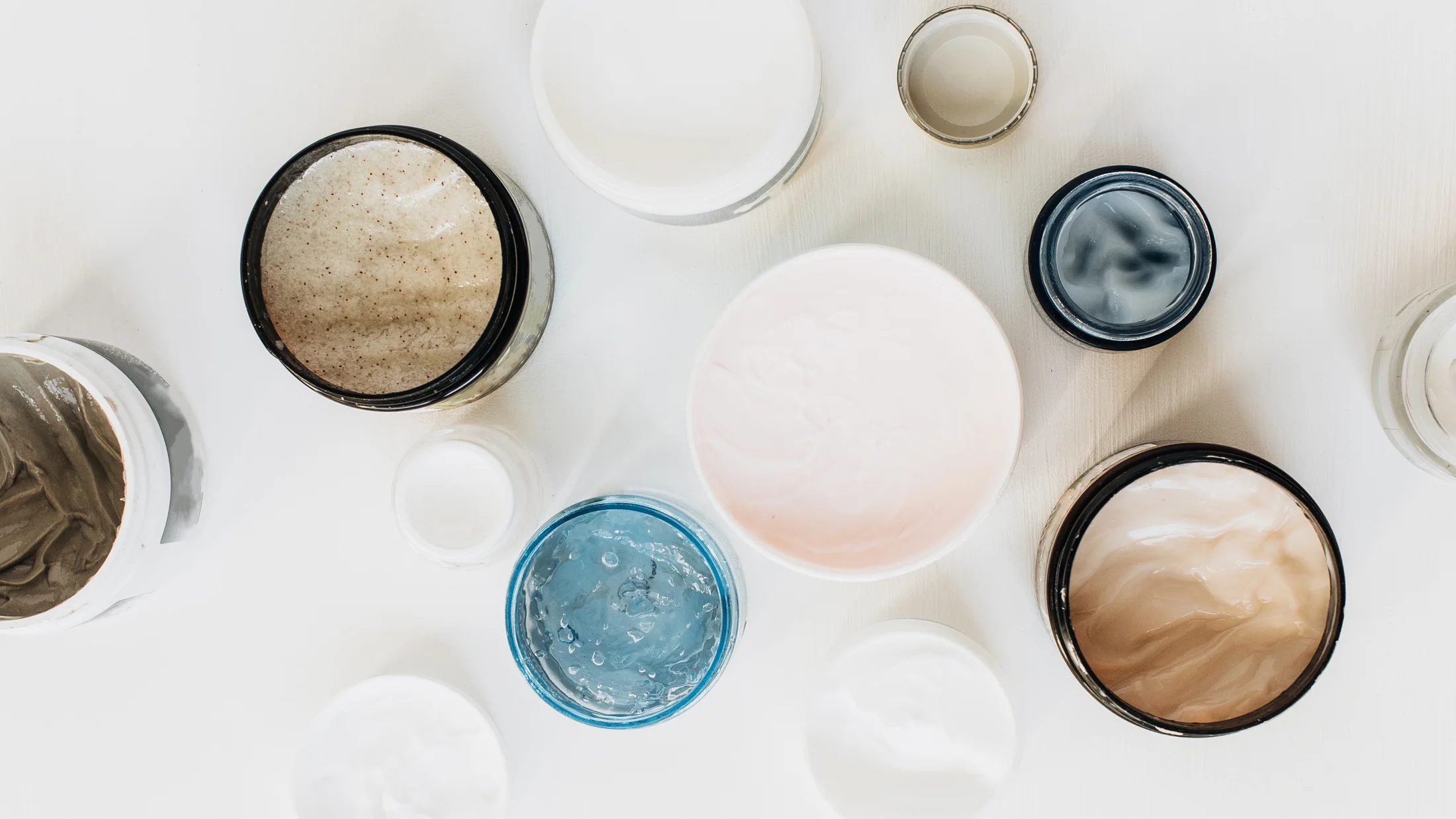 An overhead composition of cosmetic jars, creamy formulations and skincare textures on white.