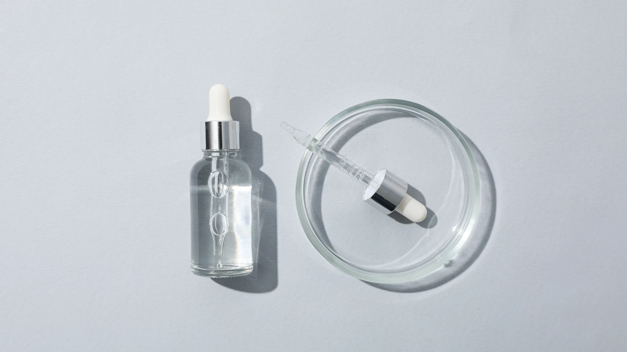 A clear skincare serum bottle beside a glass dish in a minimalist, softly lit composition.